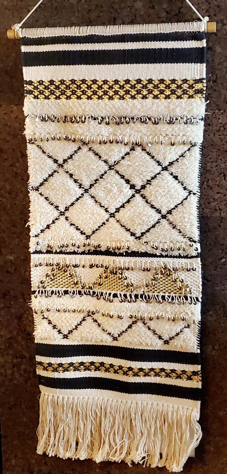 May include: A white and black woven wall hanging with a fringe border. The hanging features a geometric pattern with black diamonds on a white background. There are also gold accents throughout the design.