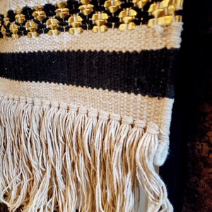 May include: Black and white woven wall hanging with a gold trim and fringe. The fringe is made of natural fibers.
