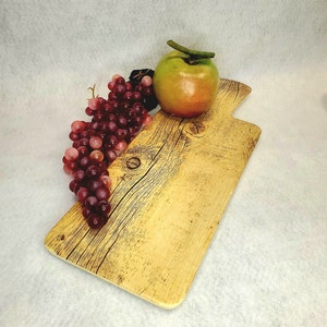 May include: A rustic wooden serving board with a handle, featuring a natural wood grain pattern. A bunch of red grapes and a green apple are placed on the board.