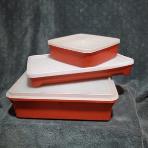 May include: Three red rectangular food storage containers with white lids are stacked on top of each other. The containers are made of plastic and have a simple, classic design. The lids fit snugly on the containers.