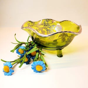 May include: A green glass footed bowl with a silver rim and floral design. The bowl is decorated with silver leaf patterns. Blue and yellow artificial flowers are placed near the bowl.