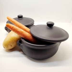 May include: A close-up shot features two small, matte black ceramic pots with lids, one partially open, revealing two orange carrots. A single potato rests beside the pot. The pots have small handles. The background is white.
