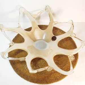 May include: A decorative, clear glass bowl with an off-white interior, featuring an open, star-like design. The bowl sits atop a round, textured, gold-colored surface. The bowl's unique shape and color create an eye-catching centerpiece.