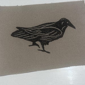 May include: A rectangular fabric patch in a muted taupe color featuring a black screen-printed graphic of a crow in profile. The bird illustration displays detailed linework on the wings and feathers, capturing the bird in a standing position.