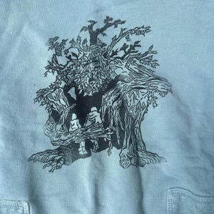 May include: Light blue sweatshirt featuring a black and gray illustration of a tree with a face and figures within its branches. The design is centered on the front of the sweatshirt, with a pocket visible at the bottom.