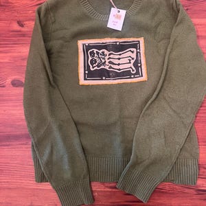 May include: An olive green long-sleeve crewneck sweater featuring a rectangular patch on the chest. The patch displays a black and white graphic of two skeletons with the text 53 and Size Sm on a small white tag attached to the collar.