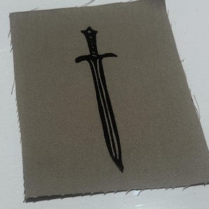 May include: A rectangular patch made of taupe fabric with raw, frayed edges. Centered on the fabric is a bold, black graphic illustration of a double-edged sword with a detailed hilt and a pointed blade, printed in a clean, minimalist style.