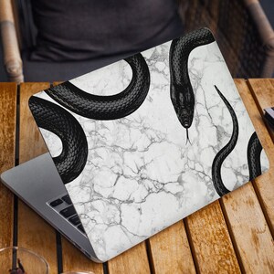 May include: A laptop computer with a white marble design and black snake illustrations. The laptop is closed and sitting on a wooden surface.