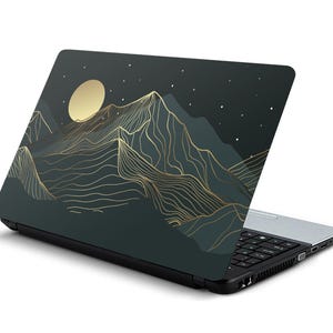 May include: A black laptop with a dark green and gold mountain design on the lid. The design features a stylised mountain range with a crescent moon and stars.
