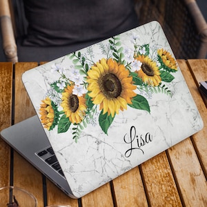May include: A white marble laptop skin featuring a bouquet of yellow sunflowers and white flowers. The name "Lisa" is written in black cursive on the skin.