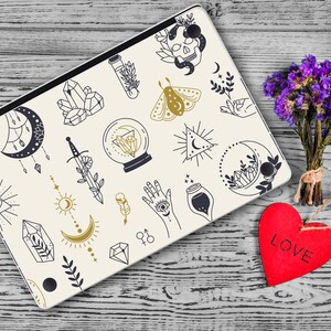 May include: A white laptop with a black and gold design featuring various symbols, including a crescent moon, a sword, a crystal ball, a moth, and a hand with an eye. The laptop is on a wooden surface with a red heart that says "LOVE" on it.