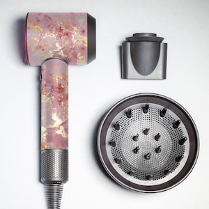 以下が含まれることがあります： ピンクとゴールドのマーブル模様のヘアドライヤーとディフューザーアタッチメント。ディフューザーは、複数の穴のあるシルバーの金属メッシュです。