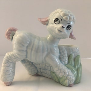 May include: A light blue ceramic planter shaped like a lamb with pink ears and a green leafy base. The lamb has a whimsical expression with large eyes and a small mouth.