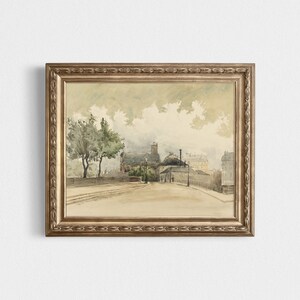 May include: A framed watercolor painting depicting a street scene with buildings, trees, and a bridge. The artwork is in a gold-colored ornate frame. The painting has a muted color palette with shades of beige, gray, and green, evoking a vintage aesthetic.
