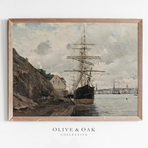 May include: A framed vintage print of a harbor scene. A large black ship is docked at a wooden pier, with a rocky cliff and buildings on the left. The sky is overcast with shades of gray and blue. The print is from the Olive & Oak Collective.