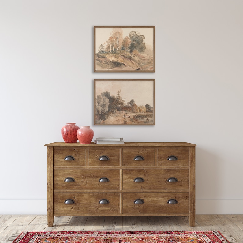 May include: Two framed watercolor landscape paintings of trees and a house, hanging on a white wall above a wooden dresser with six drawers. Two red ceramic vases are on top of the dresser.