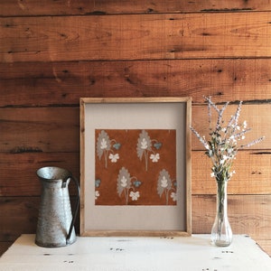 May include: A framed print with a brown background and a repeating pattern of white and blue stylized flowers. The frame is made of wood and has a light brown finish.