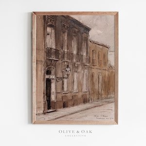 May include: A framed watercolor painting of a street scene in a European city. The buildings are painted in shades of brown and beige, and the street is empty. The painting is signed and dated in the lower right corner.