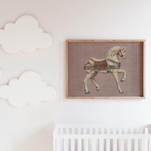 May include: A framed print featuring a vintage carousel horse illustration. The horse is depicted in cream and tan tones with a saddle in shades of red, blue, and green. The artwork is set against a muted mauve background and framed in natural wood.