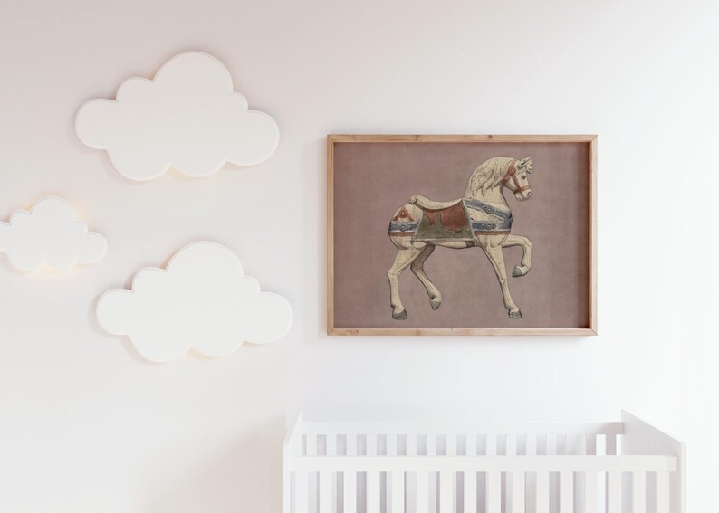 May include: A framed print featuring a vintage carousel horse illustration. The horse is depicted in cream and tan tones with a saddle in shades of red, blue, and green. The artwork is set against a muted mauve background and framed in natural wood.