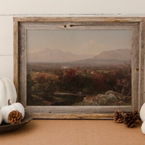 Puede incluir: Una pintura de paisaje enmarcada que representa un valle con monta&ntilde;as en el fondo. La pintura est&aacute; en un marco de madera r&uacute;stico y est&aacute; rodeada de calabazas blancas y pi&ntilde;as.