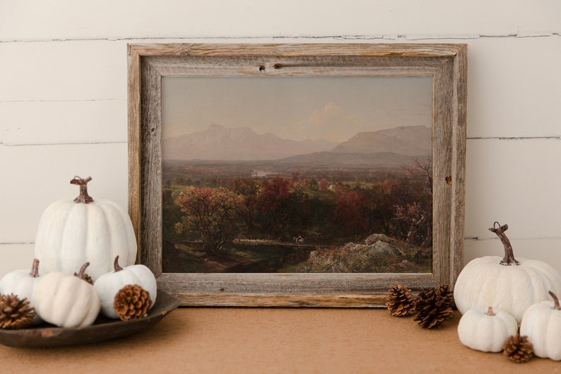 Puede incluir: Una pintura de paisaje enmarcada que representa un valle con monta&ntilde;as en el fondo. La pintura est&aacute; en un marco de madera r&uacute;stico y est&aacute; rodeada de calabazas blancas y pi&ntilde;as.