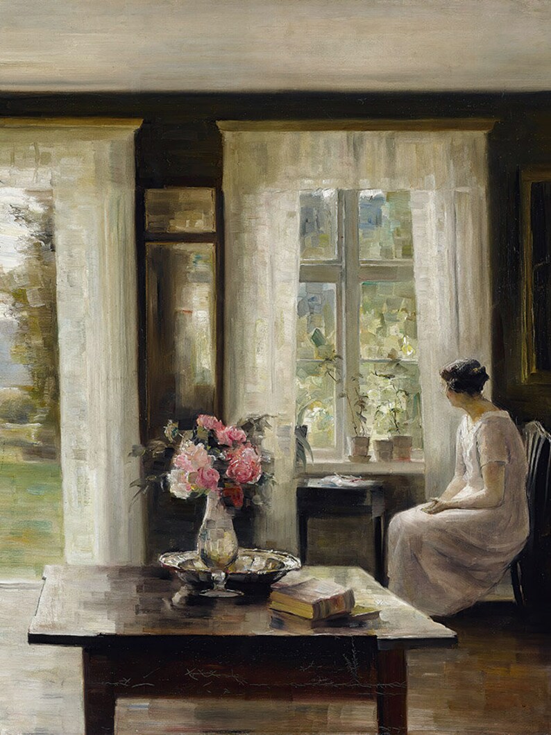 May include: A woman sits by a window in a room with white curtains. A bouquet of pink flowers in a vase sits on a wooden table with a book and a silver bowl. The window looks out onto a garden with greenery.