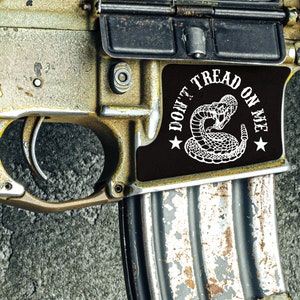 May include: A close-up of a gold-colored rifle with a black magazine and a sticker that reads "Don't Tread On Me" with a rattlesnake design.