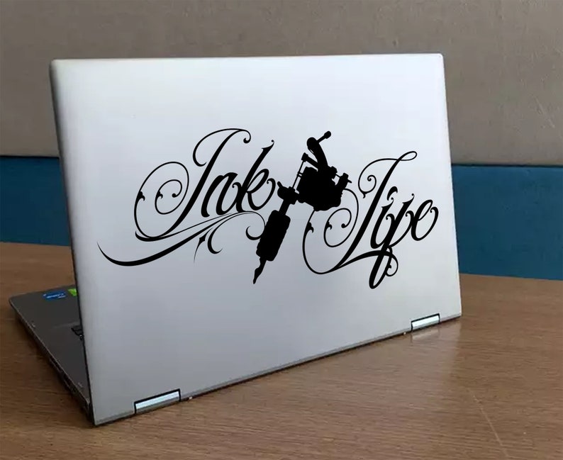 Op de afbeelding: Een witte laptop met een zwarte sticker waarop "Ink Life" staat met een gestileerd ontwerp van een tatoeagemachine.