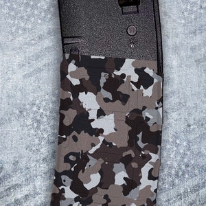 May include: A black magazine with a camouflage pattern wrap. The magazine has a black plastic body and a clear plastic window. The camouflage pattern is a mix of brown, gray, and white.