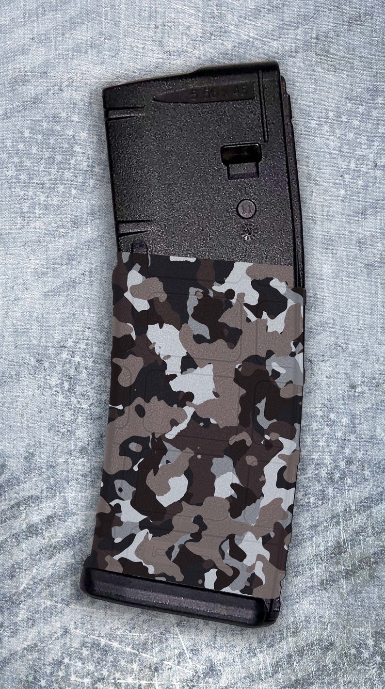 May include: A black magazine with a camouflage pattern wrap. The magazine has a black plastic body and a clear plastic window. The camouflage pattern is a mix of brown, gray, and white.