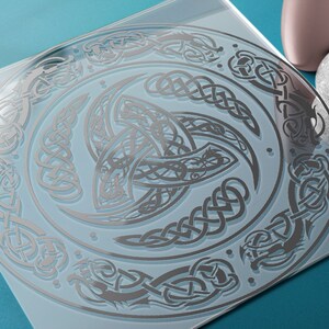 May include: A silver Celtic knot design on a clear glass surface. The design features a central triskelion with intricate knotwork and a surrounding border of dragons and other Celtic motifs.