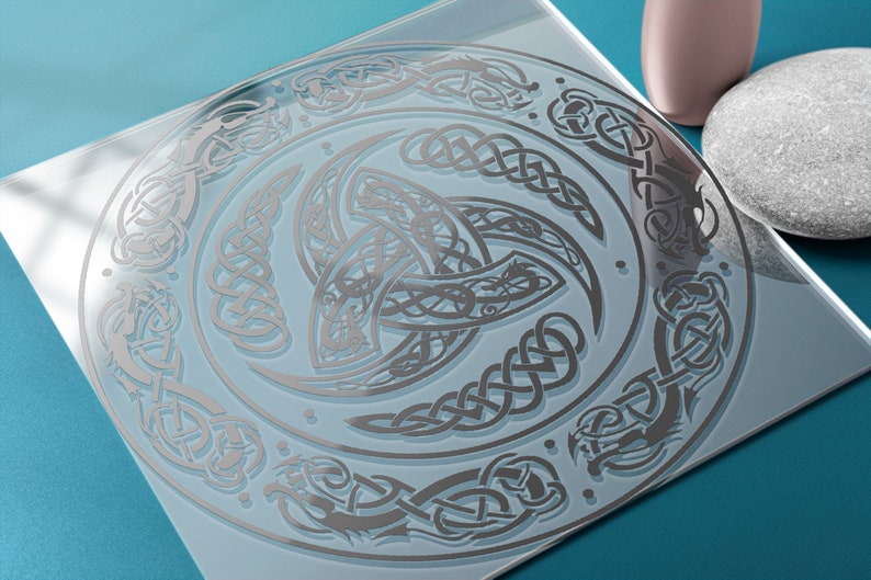 May include: A silver Celtic knot design on a clear glass surface. The design features a central triskelion with intricate knotwork and a surrounding border of dragons and other Celtic motifs.