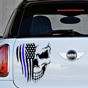 May include: A black and white skull sticker with an American flag and a blue line, representing law enforcement, on a white car. The sticker is on the back of the car, near the MINI logo.