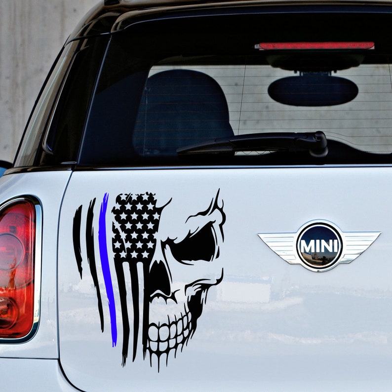 May include: A black and white skull sticker with an American flag and a blue line, representing law enforcement, on a white car. The sticker is on the back of the car, near the MINI logo.