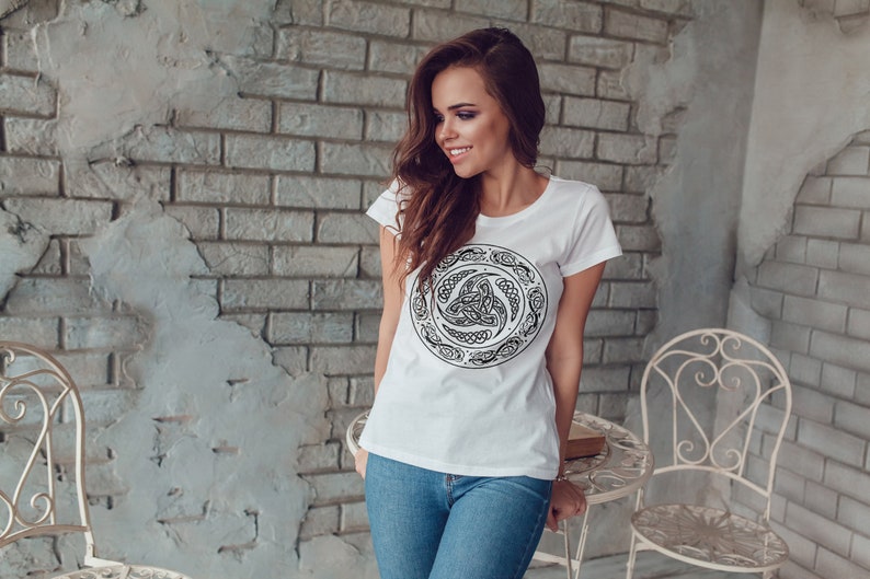 May include: A white t-shirt with a black Celtic knot design. The design is a circle with a knot in the center and a border of intricate patterns.