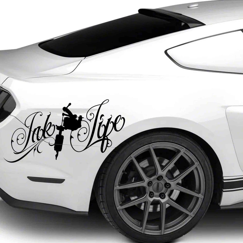 Op de afbeelding: Zwart-witte sticker met een tatoeagemachine met de tekst "Ink Life" in een gestileerd lettertype. De sticker is aangebracht op de achterkant van een witte auto.
