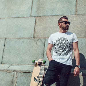 May include: A person wearing a white t-shirt with a black Celtic knot design and black pants stands in front of a stone wall. They are holding a longboard skateboard with a black and white design.