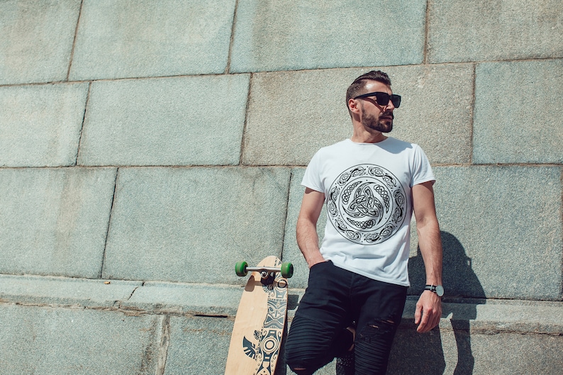 May include: A person wearing a white t-shirt with a black Celtic knot design and black pants stands in front of a stone wall. They are holding a longboard skateboard with a black and white design.