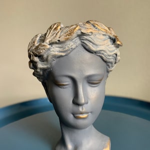 Op de afbeelding: Een grijs gipsen buste van een vrouw met een gouden bladkroon. De buste is een plantenpot met een gat bovenop om te planten.