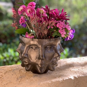 May include: A decorative planter in a metallic finish, adorned with multiple sculpted faces. The planter is filled with a colourful arrangement of pink, purple, and green flowers. A striking piece for any home, measuring approximately 20 cm in diameter.