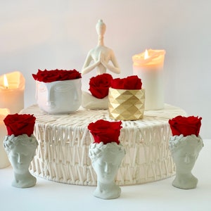 Op de afbeelding: Vijf witte keramische bustes met rode rozen erop, gerangschikt op een ronde witte rieten basis. Op de achtergrond staat een wit keramiek figuur in een yogapose met twee witte kaarsen en een gouden geometrische vaas met een rode roos.