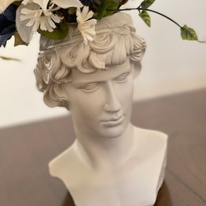Puede incluir: Una maceta con forma de busto de acabado blanco mate que representa una cabeza masculina clásica con cabello rizado. La cabeza sirve como recipiente para un arreglo de flores artificiales, incluyendo margaritas blancas, flores azul oscuro y tallos verdes.