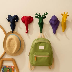 May include: A selection of animal-shaped wall hooks in various colours. The hooks depict a ram, elephant, deer, gorilla, and giraffe. A straw hat and a green backpack are suspended from the hooks. The hooks are fixed to a white wall.