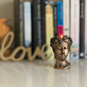 May include: A bronze-coloured candle in the shape of a classical bust, crowned with a laurel wreath. The candle is lit, with a small flame at the top. The background features a blurred wooden "love" sign and a bookshelf.