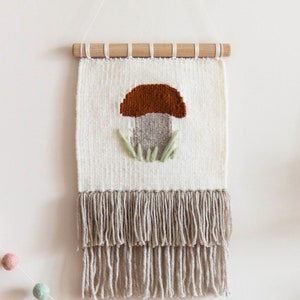 May include: A wooden shelf with a crescent moon-shaped wooden sign that says "pure magic" and a white ceramic arch-shaped object. There is a woven wall hanging with a mushroom design and a string of colorful pom-poms.