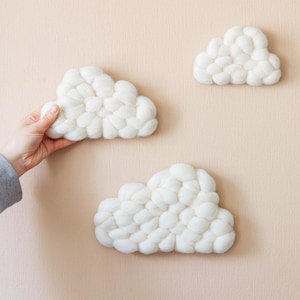May include: Three white cloud-shaped wall decorations made of soft, interconnected felted wool spheres. The clouds vary in size and are arranged in a cluster, featuring a textured, rounded surface that mimics the appearance of fluffy cumulus clouds.