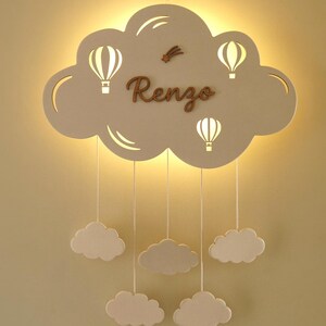 May include: A wall-mounted cloud-shaped light fitting featuring the name Renzo in brown script, three cut-out hot air balloon silhouettes, and a small shooting star. Five smaller cloud shapes hang from the main cloud by white strings.