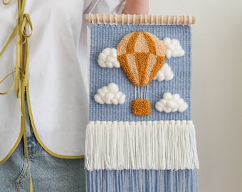 May include: A woven wall hanging with a hot air balloon design. The balloon is orange and tan, set against a blue woven background with white fluffy clouds. The bottom has white and blue fringe, and a wooden dowel at the top.
