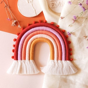 May include: A rainbow-coloured macrame wall hanging with a white fringe. The rainbow is made of yarn in shades of red, orange, yellow, pink, and purple. The hanging is on a light pink and orange background.